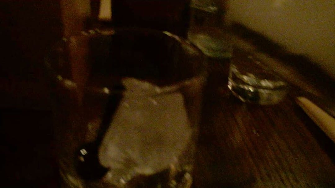 Close-up of an empty glass with a black stirrer and a large ice cube inside, placed on a dimly lit wooden table with other glassware in the background. A pair of cute cockatoos perches nearby, adding charm to the cozy scene.